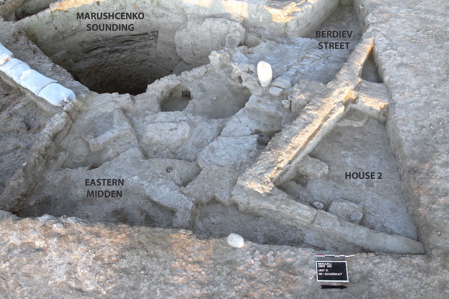 Überblick über die Architektur und Befunde in Unit D am Ende der 2011 Kampagne. Der gepflasterte Teil von Berdiev Street endet an einem Tor, Gate 1, das mit einem stehenden Steinblock markiert ist. Hinter dem Tor befindet sich der Eastern Midden. Haus 2 befindet sich direkt am nördlichen Straßenrand. Der von A. Marushchenko 1959 angelegte Tiefschnitt schneidet den Midden sowie die Architektur im Süden (im Bild oben links)